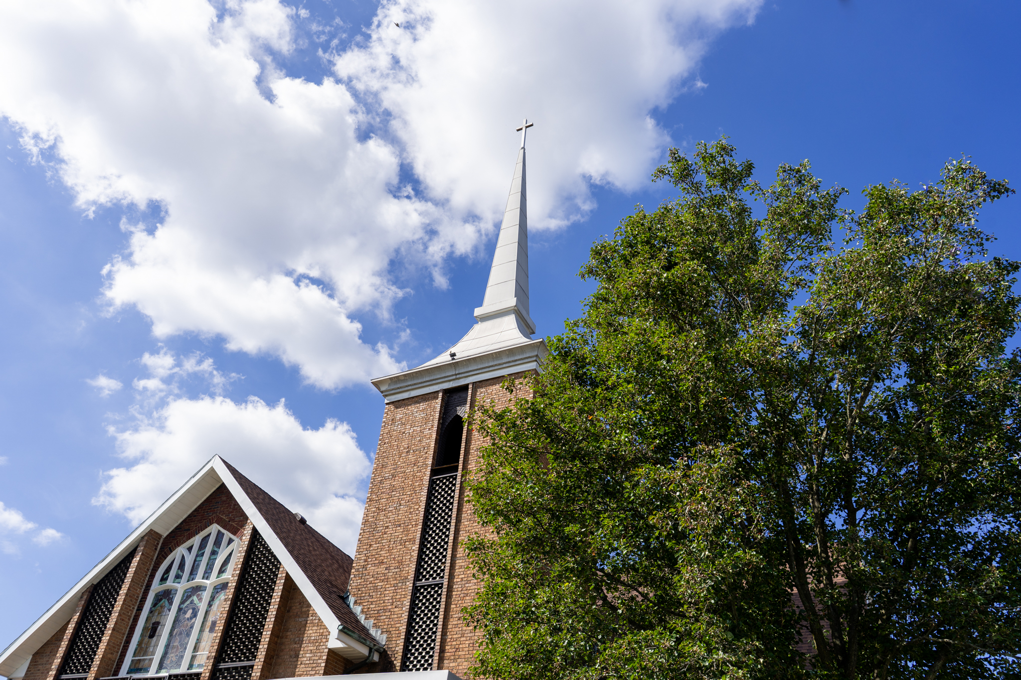 The front of the church focused on the steeple with a cross with a tree in the front and clouds behind the steeple.