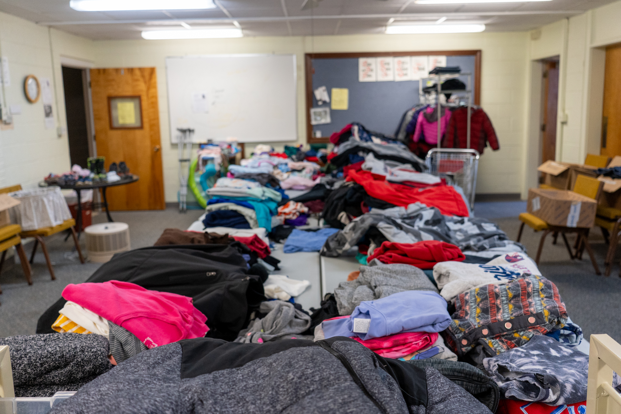 Four large tables covered with stacks of folded clothing items with additional items in the background including a rack of coats and shoes.