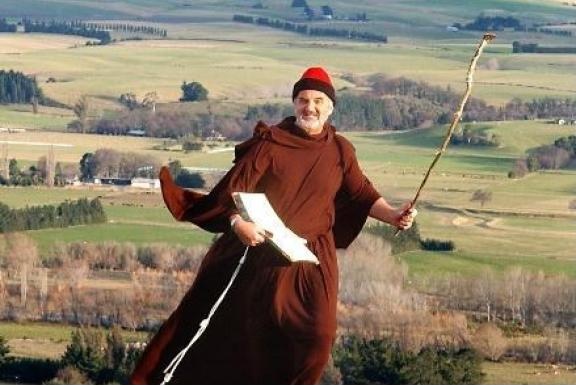 A man wearing a brown robe, holding a stick and a book, standing over fields and trees.
