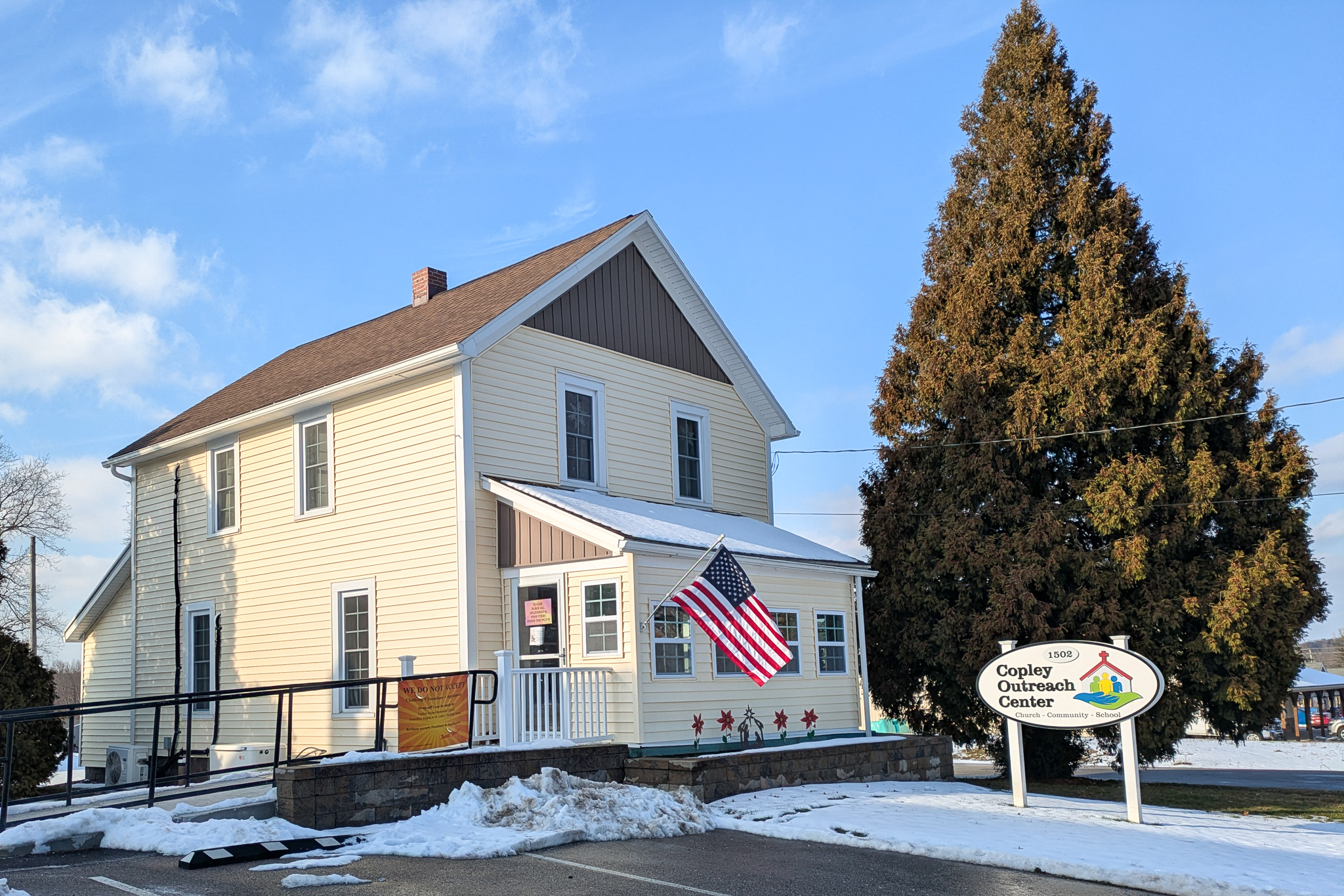 A small building with yellow siding, a brown roof, next to a large pine tree.
